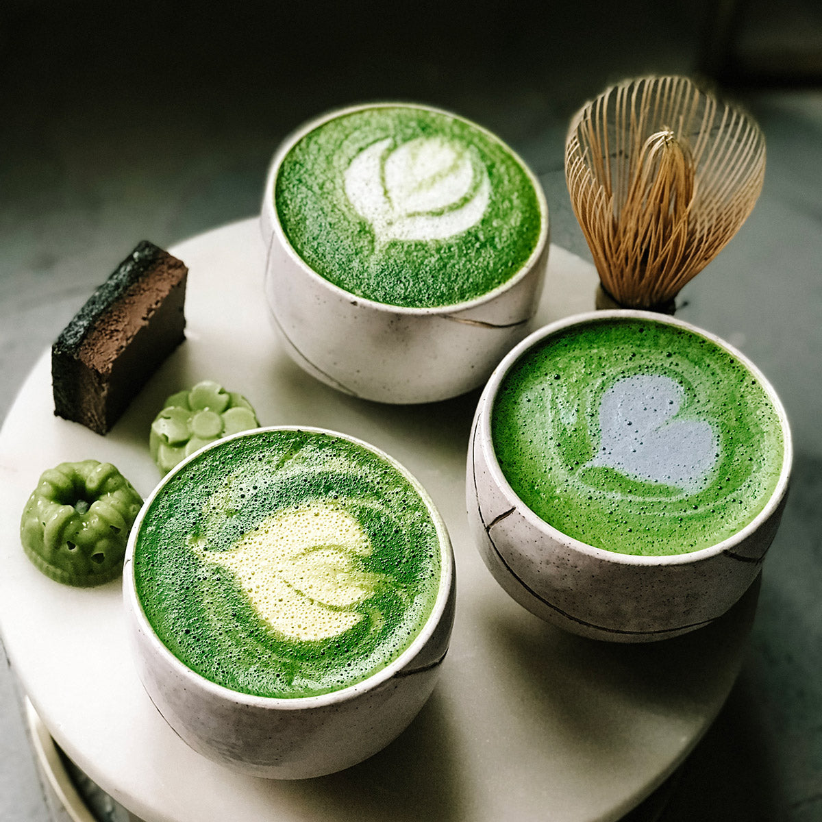 Arrangement of three matcha lattes with foam art, bamboo whisk, wagashi, and brownie on a marble pedestal