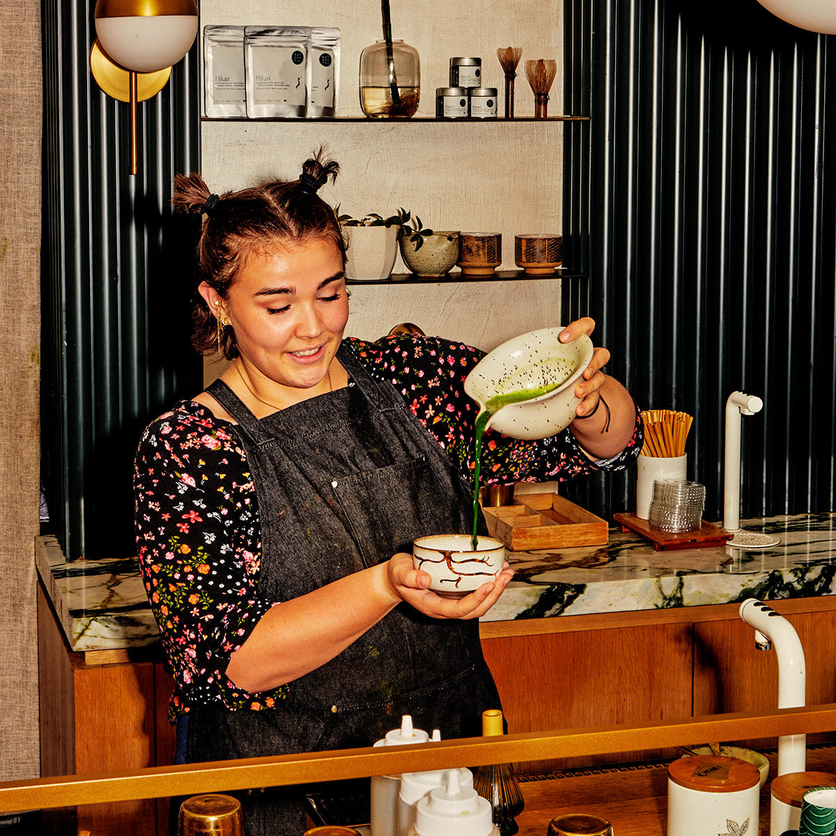 Interior of Nolita café, barista pouring matcha from a speckled white ceramic whisking bowl into a ceramic tea bowl with golden glaze in a root-like pattern