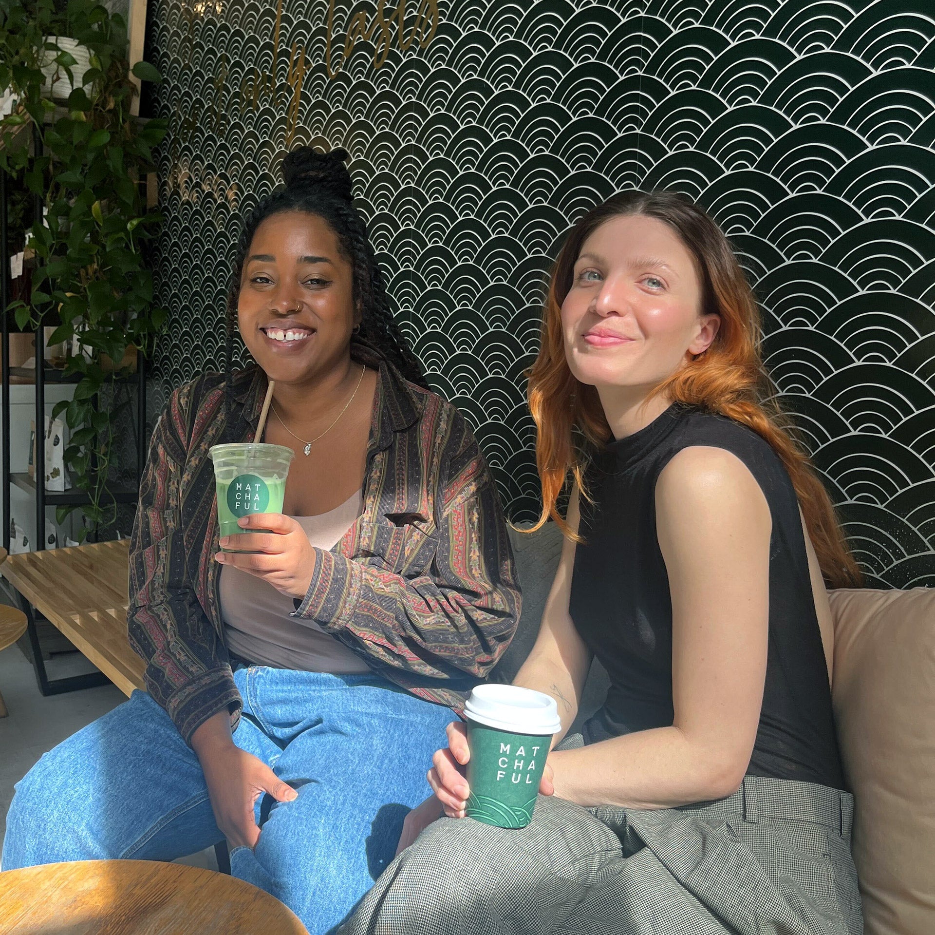 Jordan and Elise drinking matcha lattes and sitting on a bench in the Matchaful Café with a dark green seigaiha patterned wallpaper behind them