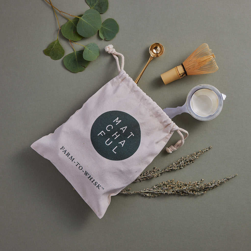 Matchaful branded drawstring bag with a gold scoop, bamboo whisk, and stainless steel matcha sifter coming out of the bag